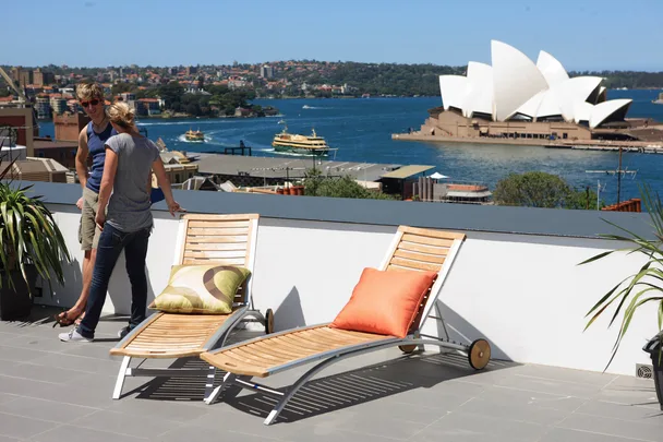 Two people talking on a rooftop terrace overlooking Sydney Opera House with deck chairs in the foreground.
