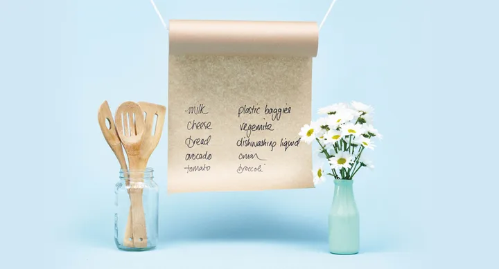A kitchen list on a hanging scroll between a jar of wooden utensils and a vase of white flowers against a blue background.