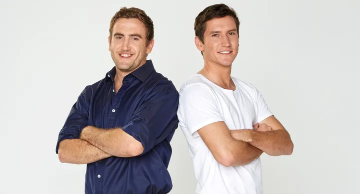Two smiling men standing back-to-back, arms crossed, one in a navy shirt, the other in a white t-shirt, on a white background.