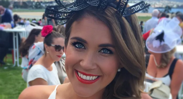 Woman smiling at a racecourse event, wearing a black decorative headpiece and red lipstick, with crowd in background.