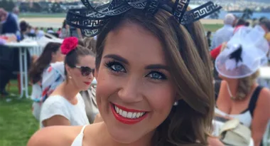 Woman smiling at a racecourse event, wearing a black decorative headpiece and red lipstick, with crowd in background.