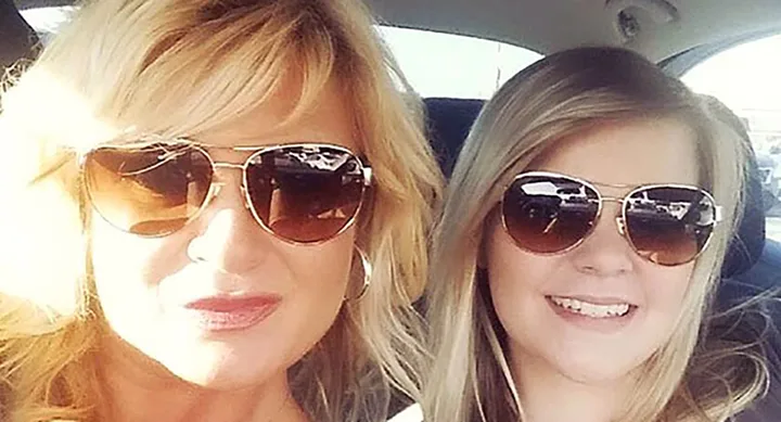 Two women with blonde hair and sunglasses smile while sitting inside a car.