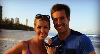A couple smiling on a beach, the woman shows an engagement ring, with city buildings and ocean in the background.