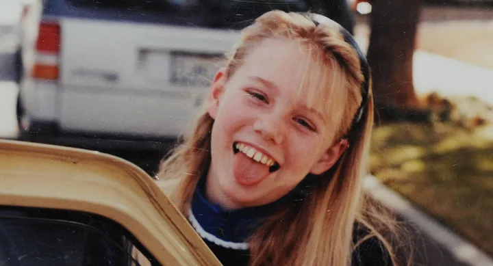 A young girl with blonde hair smiles and sticks out her tongue next to a car, with a street scene in the background.