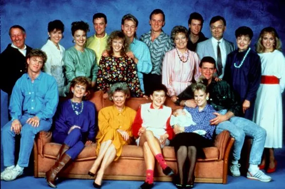 Cast of "Neighbours" TV show posing together in colorful 1980s attire.