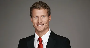 Man in a suit with a red tie, smiling against a gray background.