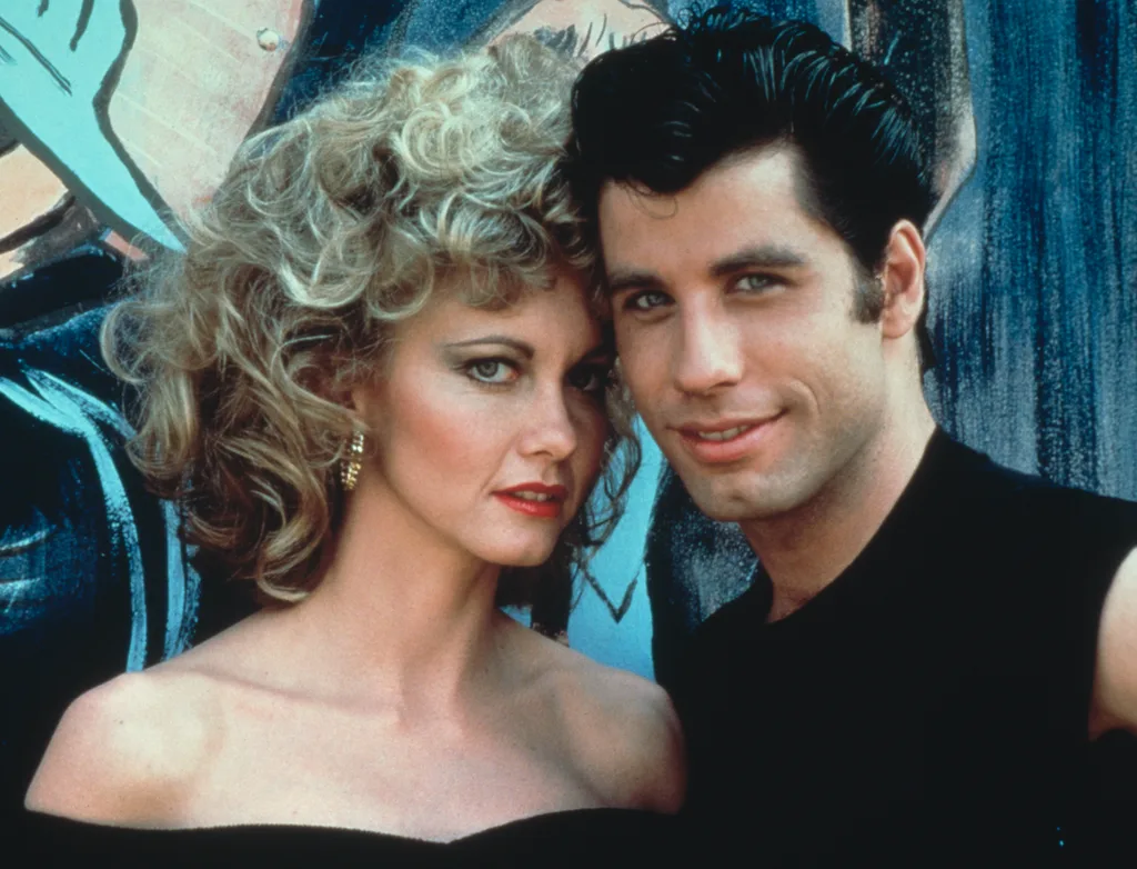 A couple poses closely together, both with 1950s-style hair and clothing, set against a painted mural backdrop.