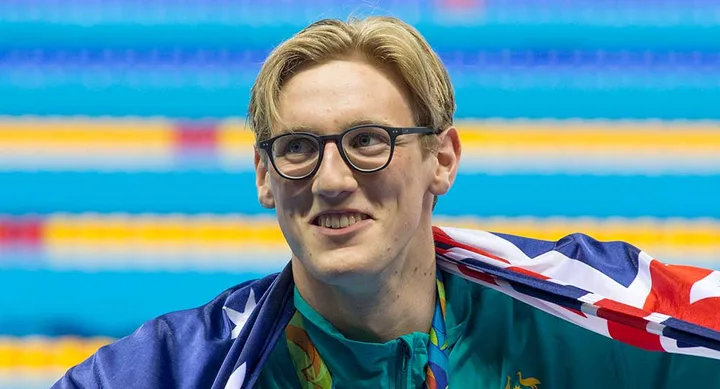 A swimmer with glasses and an Australian flag draped over shoulders smiles at a poolside venue.