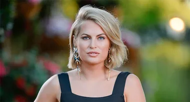 ```plaintext
Woman with short blonde hair wearing a black dress and earrings, standing outdoors with a blurred background.
```