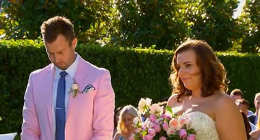 A man in a pink suit stands next to a woman in a white dress holding pink flowers at an outdoor ceremony.