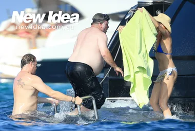 Three people in swimwear on a yacht ladder; one covering with a towel. Water and boats in the background.