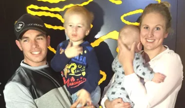 Family portrait with a man holding a toddler and a woman holding a baby, all smiling, against a decorative background.