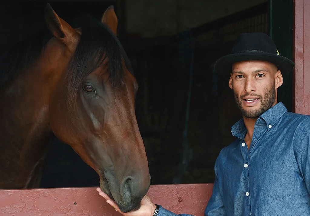 Josh Gibson, wearing a blue shirt and hat, patting a horse in a stable