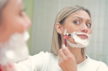 Woman shaving her face in the mirror with shaving cream and a razor