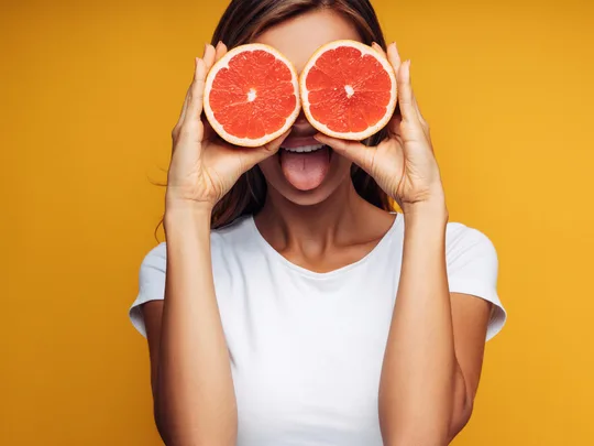 Woman holding two halves of grapefruits up to her eyes while standing against orange backgrounnd