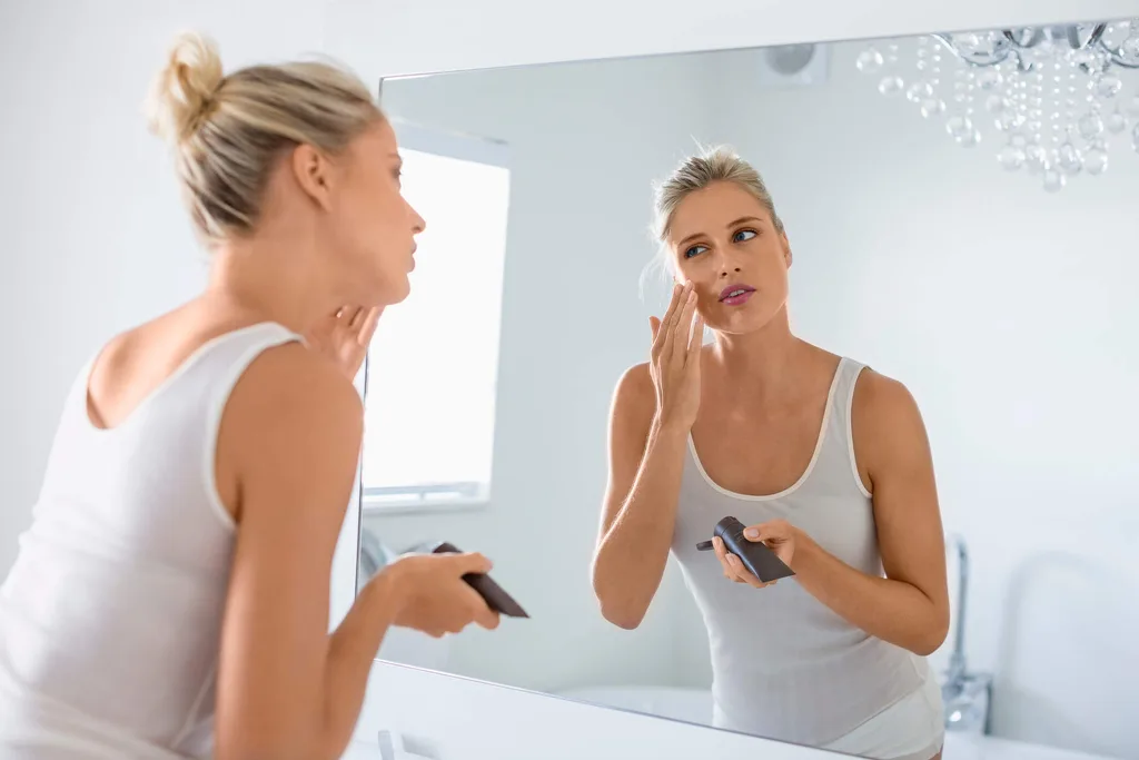 Woman applying moisturizer to face in the mirror