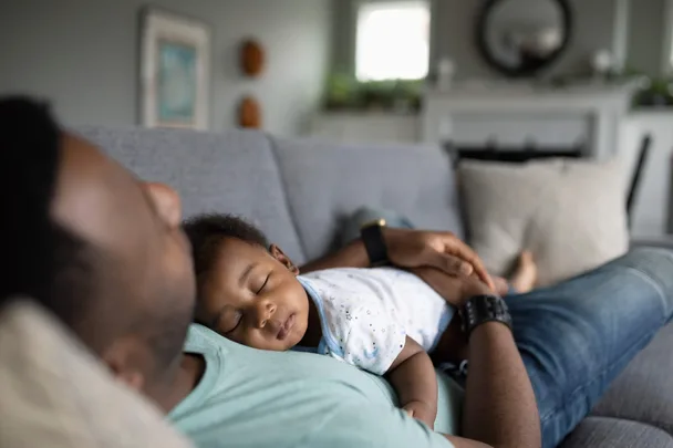 A baby lying on a father's chest, while he lays in the couch
