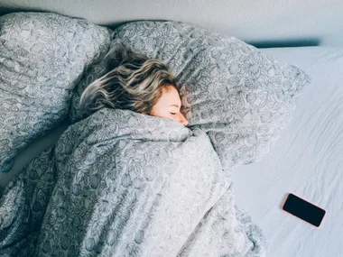 A woman tucked in bed sleeping with her phone next to her