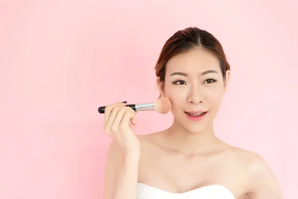 A young woman applying makeup in front of a pink background