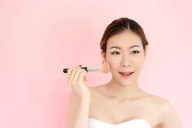 A young woman applying makeup in front of a pink background