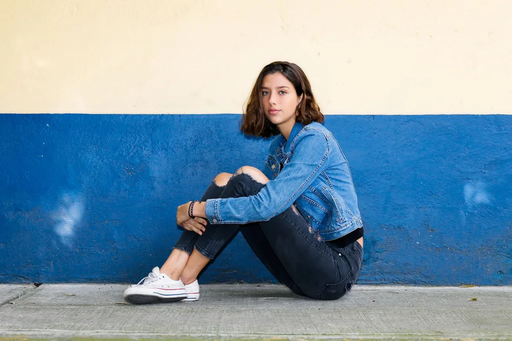 A young girl sitting on the pavement with denim jeans and a jacket