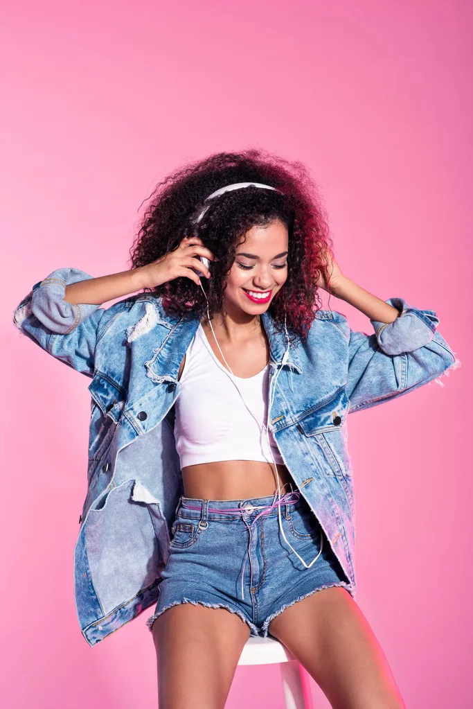 A young woman with a denim jacket, denim shorts and a white top against a pink background