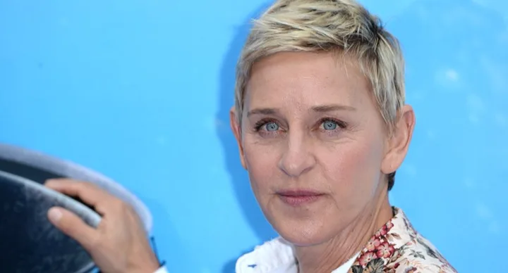 A person with short blond hair and a floral shirt stands against a blue background, not wearing a wedding ring.