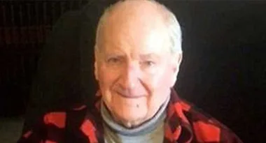 Elderly man in a red plaid shirt smiling against a dark background.