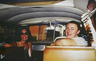 A man and woman are seated in a van, with the woman wearing sunglasses and looking forward, while the man looks back and smiles.