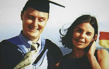 A man in a graduation cap and gown stands smiling next to a woman with shoulder-length dark hair.