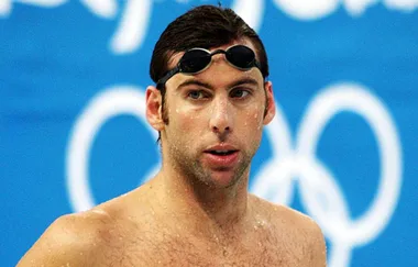 A swimmer with goggles on forehead, wet from pool with Olympic rings in background.