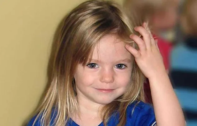 Young child with blonde hair in blue shirt smiling and touching her head, with blurred background.