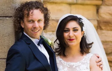 A couple in wedding attire pose together, smiling against a stone wall backdrop.