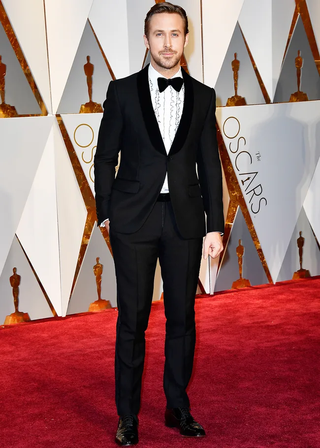 Man in a black tuxedo on the Oscars red carpet, standing confidently in front of the backdrop with Oscar statues.
