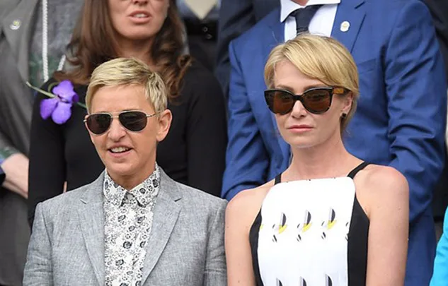 Two individuals wearing sunglasses, one in a grey suit and the other in a black and white dress, seated in a crowd.
