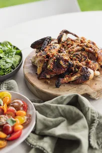 Grilled chicken with seasoning on a wooden board, with fresh salad and tomato slices on a table.
