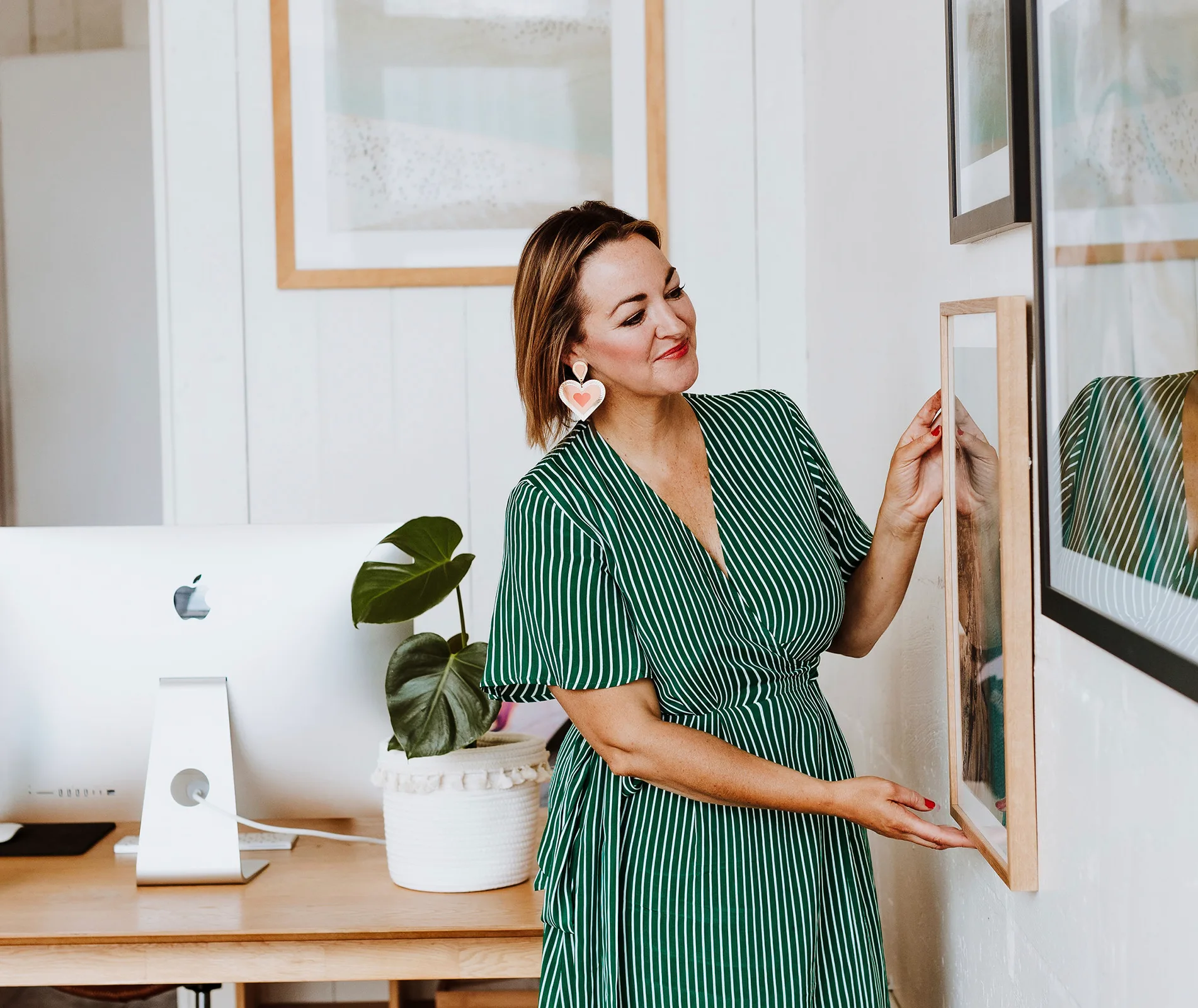 Woman in a green dress hangs a picture on a wall in a stylish office with a white computer.