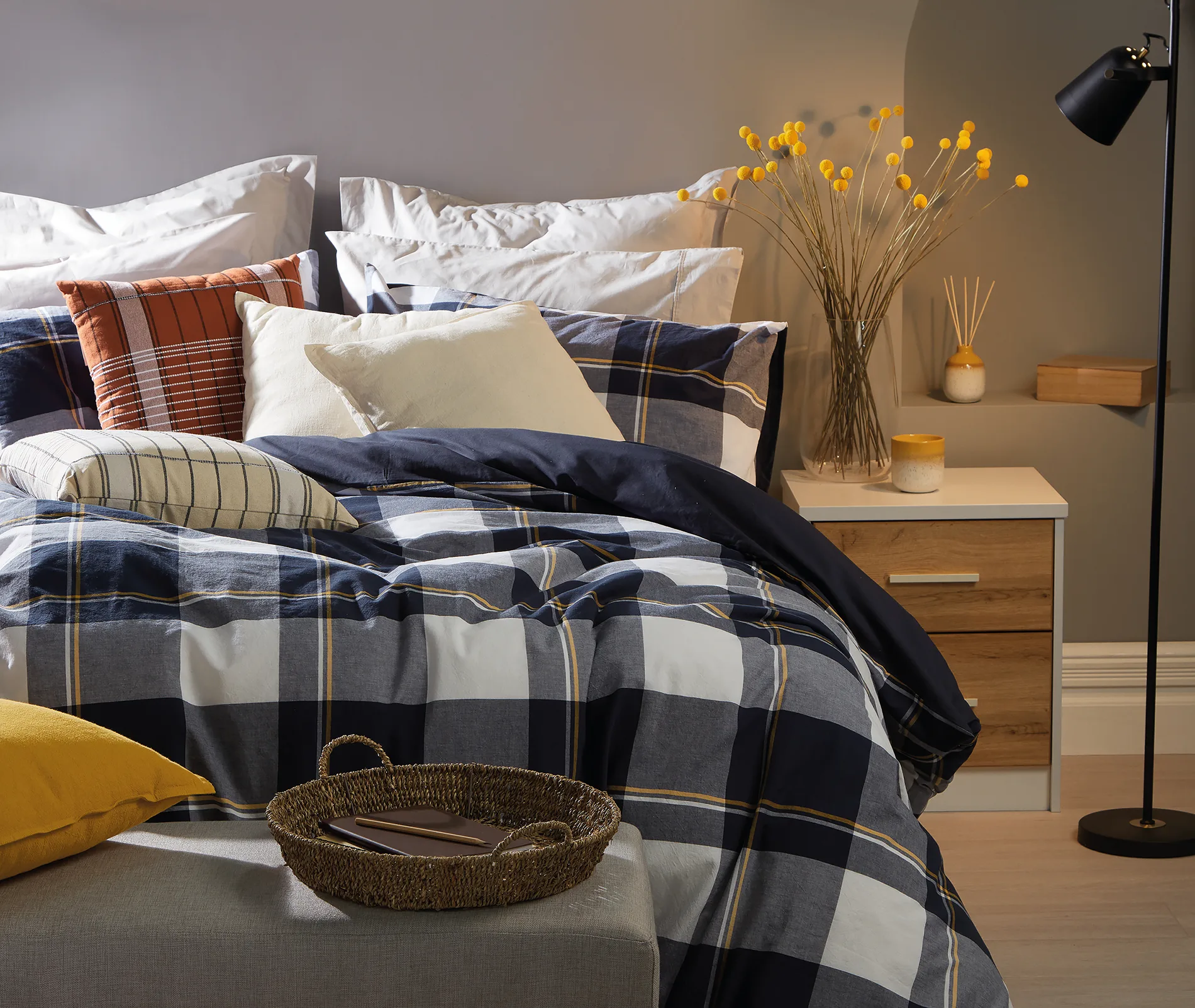 Bedroom scene with plaid bedding, assorted pillows, a nightstand with decor and vase, and a woven basket on an ottoman.