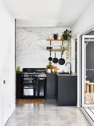 Minimalist kitchen with black oven, shelves with plants, dishes, and pans on a white brick wall.