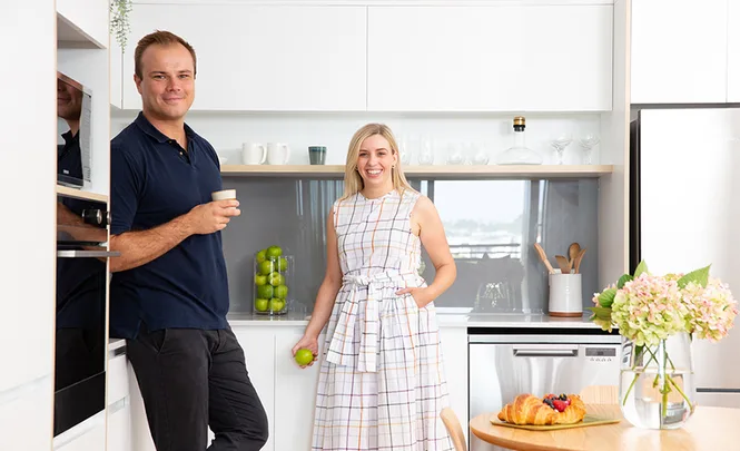 Couple standing in a modern kitchen, man holding a cup, woman holding an apple, with a table of flowers and pastries.
