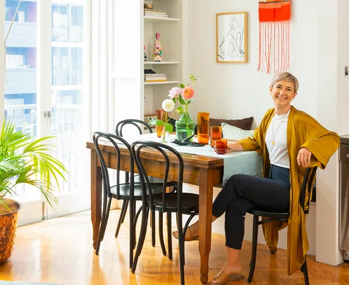 Person seated at a wooden dining table with flowers, wearing a mustard shawl, and smiling in a bright, cozy room.