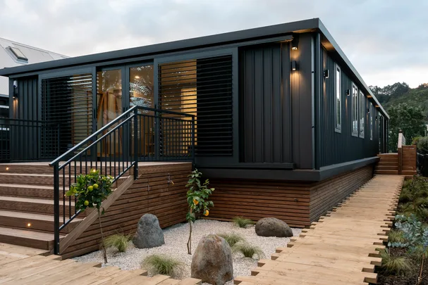 Modern black shipping container home with wooden deck, stairs, and landscaped garden with stones and plants.