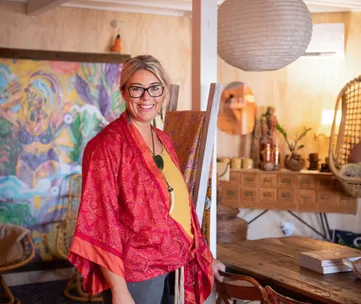 Smiling woman in a red kimono stands in a colorful, artistic room with paintings and rustic furniture.