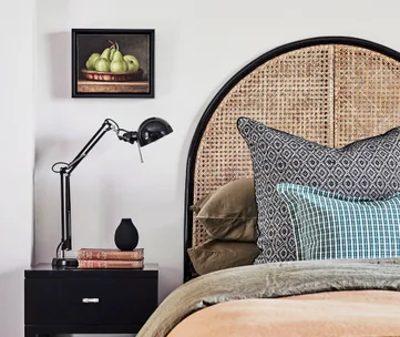 Bedroom with a rattan headboard, black lamp on a nightstand, and a painting of pears on the wall.