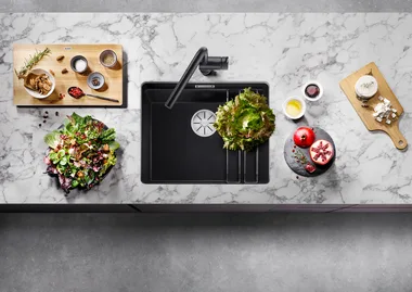 Top-down view of a kitchen countertop with a sink, salad ingredients, spices, pomegranate, and cheese on a marble surface.