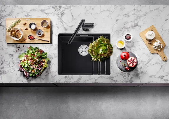 Top-down view of a kitchen countertop with a sink, salad ingredients, spices, pomegranate, and cheese on a marble surface.