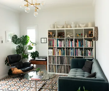 Living room with a bookshelf, plants, a black leather chair, glass coffee table, and a grey couch.