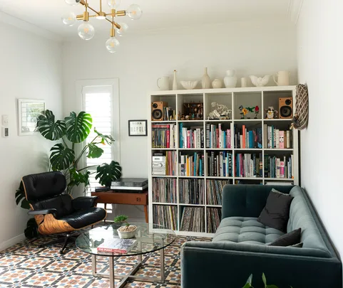 Living room with a bookshelf, plants, a black leather chair, glass coffee table, and a grey couch.