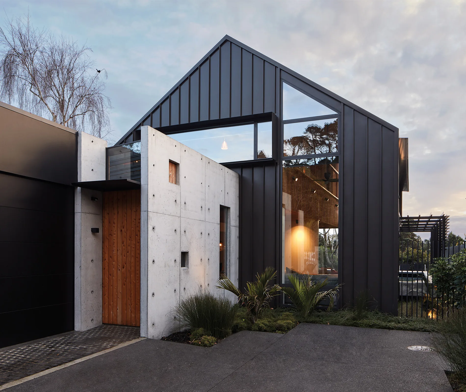 Modern house exterior with concrete, wood, and large glass windows reflecting sky and trees, surrounded by greenery.