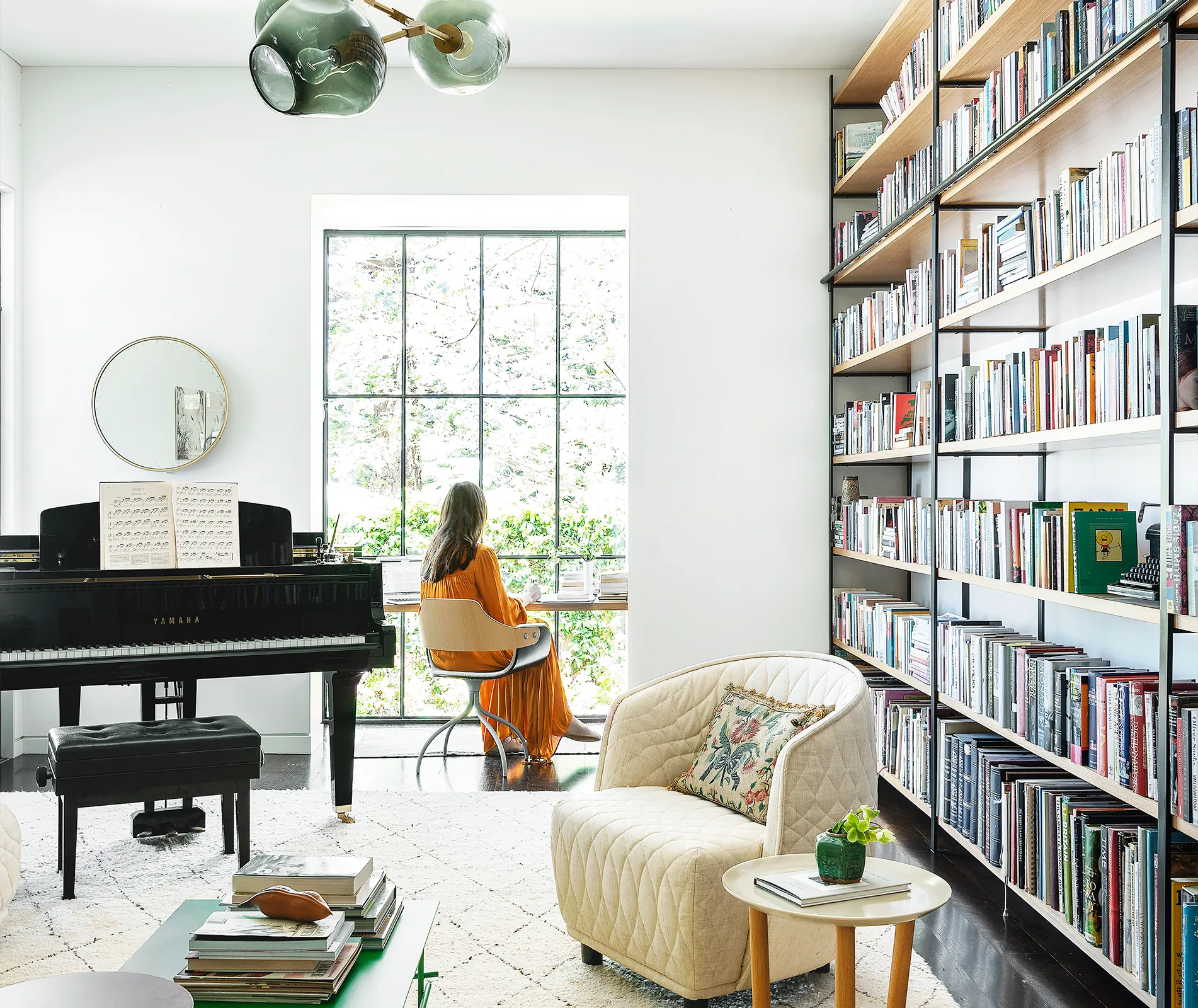 Woman in orange dress sits by a window in a bright library with bookshelves and a Yamaha piano.